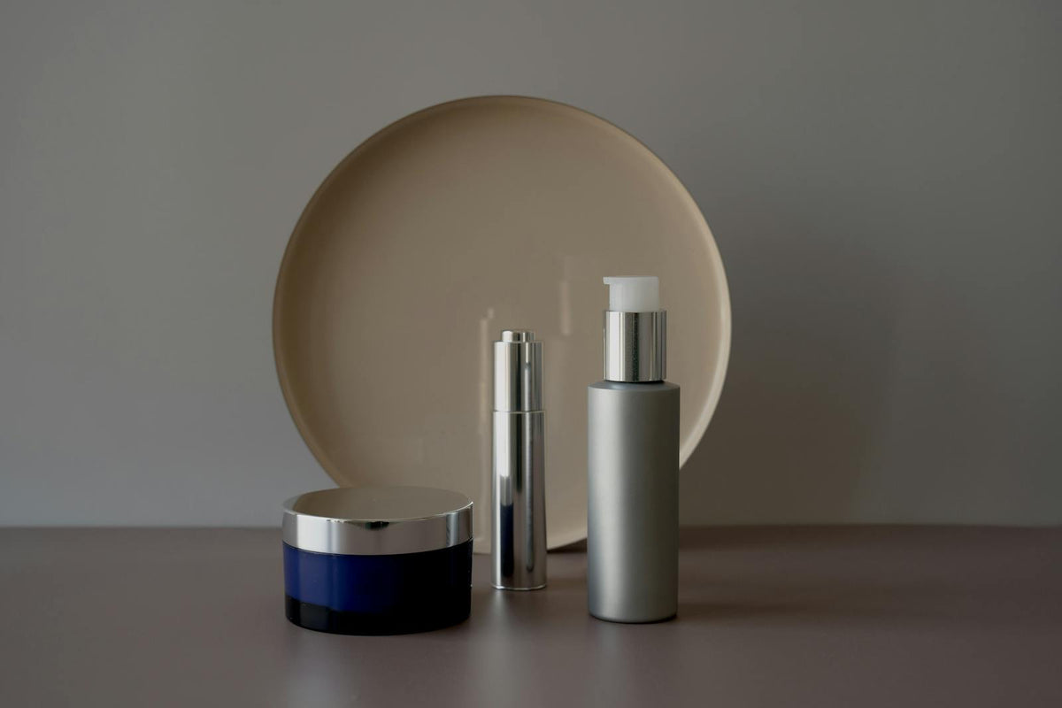 Trois soins cosmétiques (pot de crème, sérum et flacon) posés sur un fond neutre et épuré, illustration d'un guide pour lire la formule d'un cosmétique au CBD et bien le choisir.